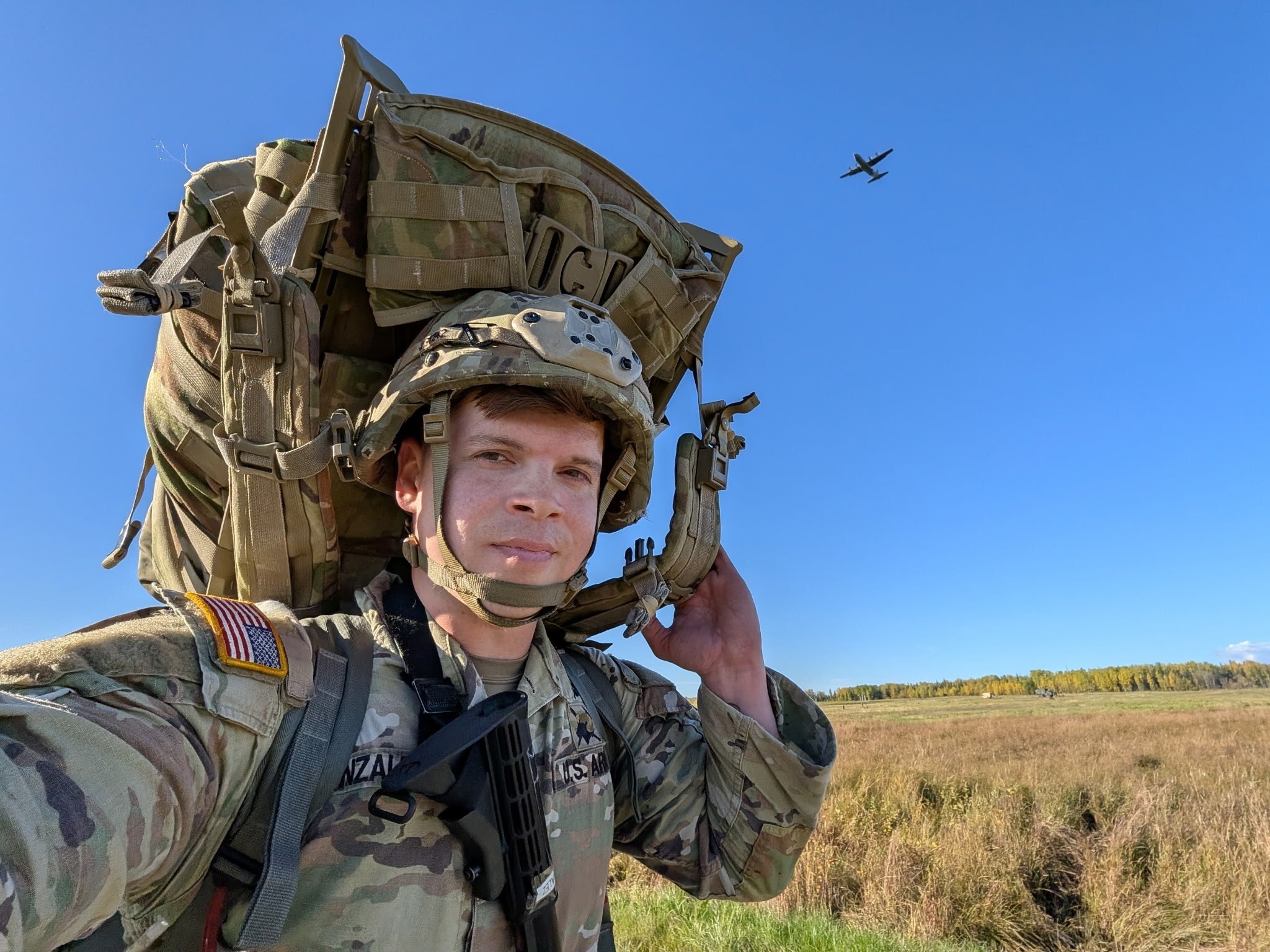 Staff Sgt. Joel Gonzalez in helmet with his ruck on his shoulder, a C-130 overhead